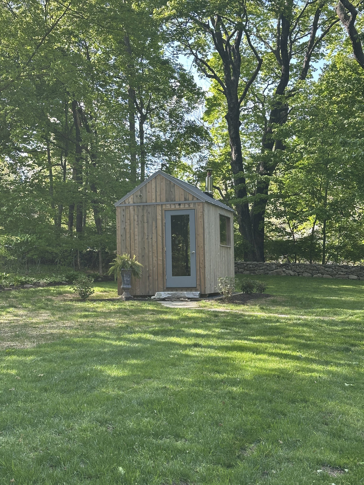 Wilton, Connecticut custom sauna — front elevation, vertical cedar cladding, glass door, and stainless chimney at the edge of the woods
